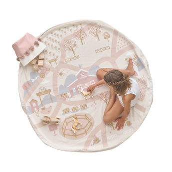 A child playing on a round playmat that features an illustrated ranch theme with trees, houses, and a road. Toys and a storage bag are also visible on the mat. Alt Img