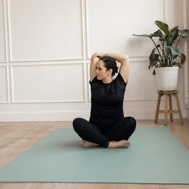 Woman practicing yoga on a green Evo Mat in a room with a plant and wooden stool.