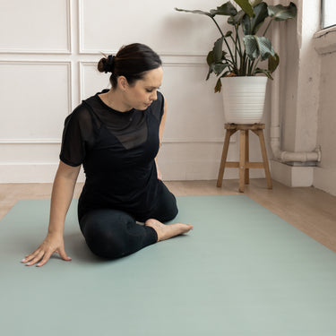 Woman in black outfit practicing yoga on a green Evo Mat with a plant in the background