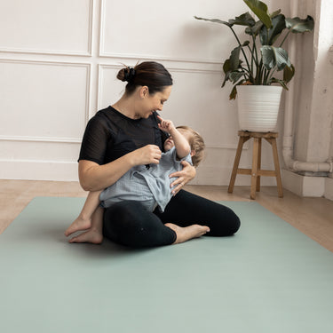 Woman embracing and playing with a child on a green Evo Mat in a room with a plant and wooden stool.