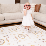 Baby toddler standing in a living room wearing heart sunglasses and enjoying a treat on the toddlekind baby toddler foam modern designer puzzle play mat haven amira golden clay.