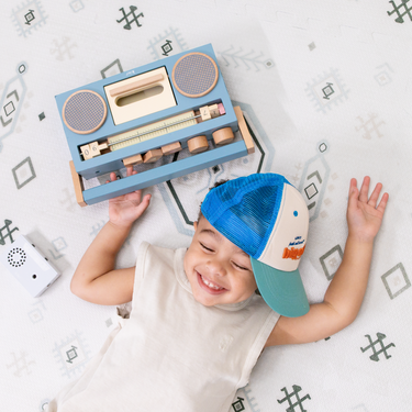Overhead view of baby toddler lying down with a toy radio, enjoying the toddlekind baby toddler foam modern designer puzzle play mat haven amira seagrass