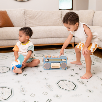Two baby toddlers playing with a toy radio on a soft surface, featuring the toddlekind baby toddler foam modern designer puzzle play mat haven amira seagrass.