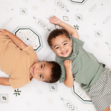 Two baby toddlers lying side by side and smiling on the soft surface of the toddlekind baby toddler foam modern designer puzzle play mat haven amira seagrass.