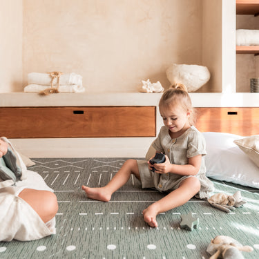 Toddler sitting and smiling while holding toy on toddlekind baby toddler foam modern designer puzzle play mat haven berber moss in a cozy home setting.