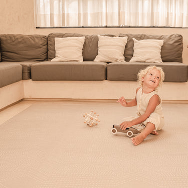 Toddler sitting and smiling with toys on the toddlekind baby toddler foam modern designer puzzle play mat haven deco latte in a bright living room.