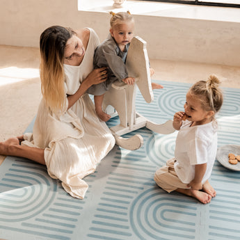 Mother helping baby on a wooden rocking toy while another toddler eats a snack on the toddlekind baby toddler foam modern designer puzzle play mat haven linear mineral.
