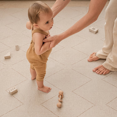 Baby being gently supported while standing on the toddlekind baby toddler foam modern designer puzzle play mat haven tulum bone with toys nearby.