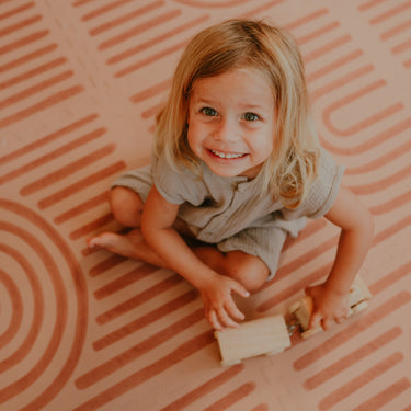 Smiling toddler playing with a wooden toy while sitting on the toddlekind baby toddler foam modern designer puzzle play mat haven linear camel.