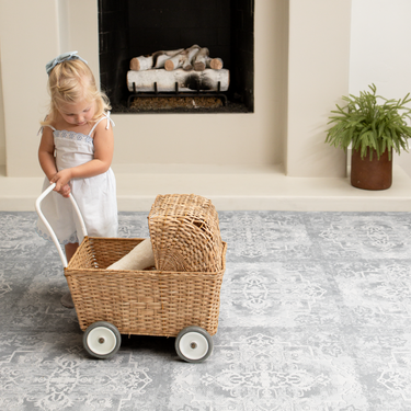 Toddler pulling a wicker cart across the patterned foam mat in a living room setting, showing durability of the toddlekind baby toddler foam modern designer puzzle play mat haven parisa pearl blue.