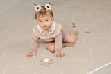 A baby sits on a soft beige interlocking foam play mat with a subtle geometric pattern, playing with wooden toy cars and a flowertoy. The cushioned mat provides a comfortable, safe surface for crawling and early play.